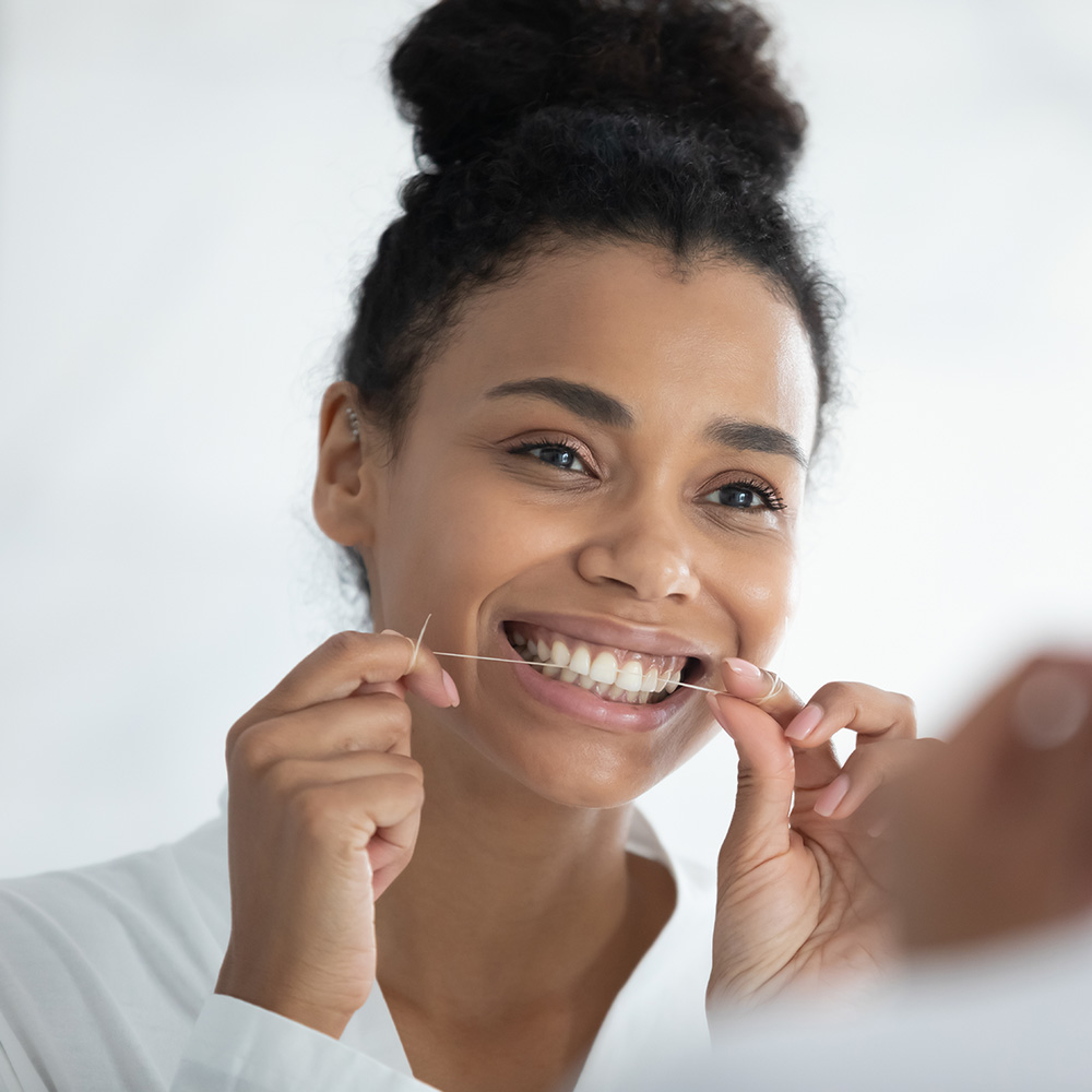 The image shows a woman smiling at her reflection in a mirror while she brushes her teeth with a toothbrush, with a focus on dental hygiene.